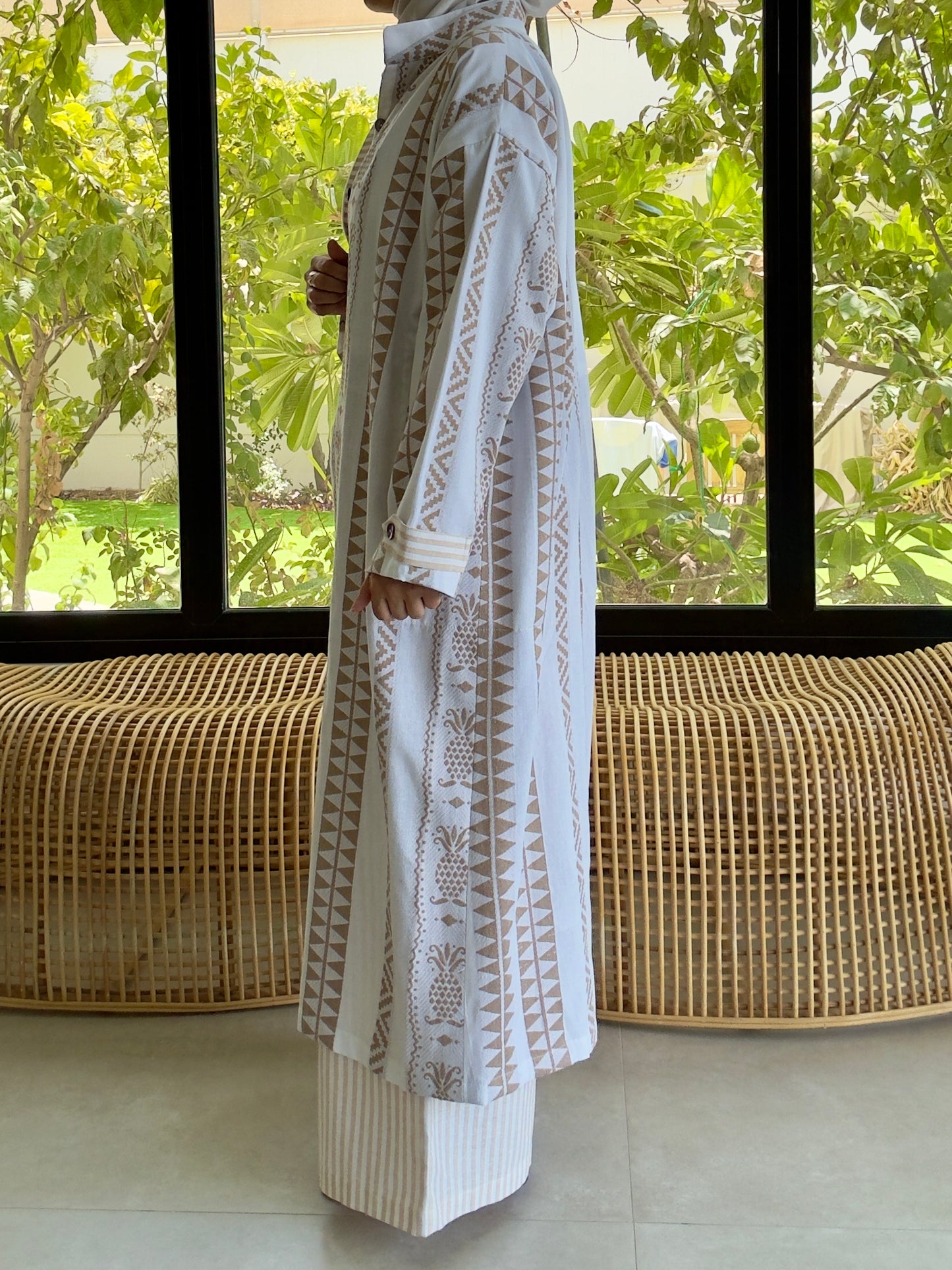 Model in white linen outfit with detailed embroidery, indoors near large windows with greenery outside