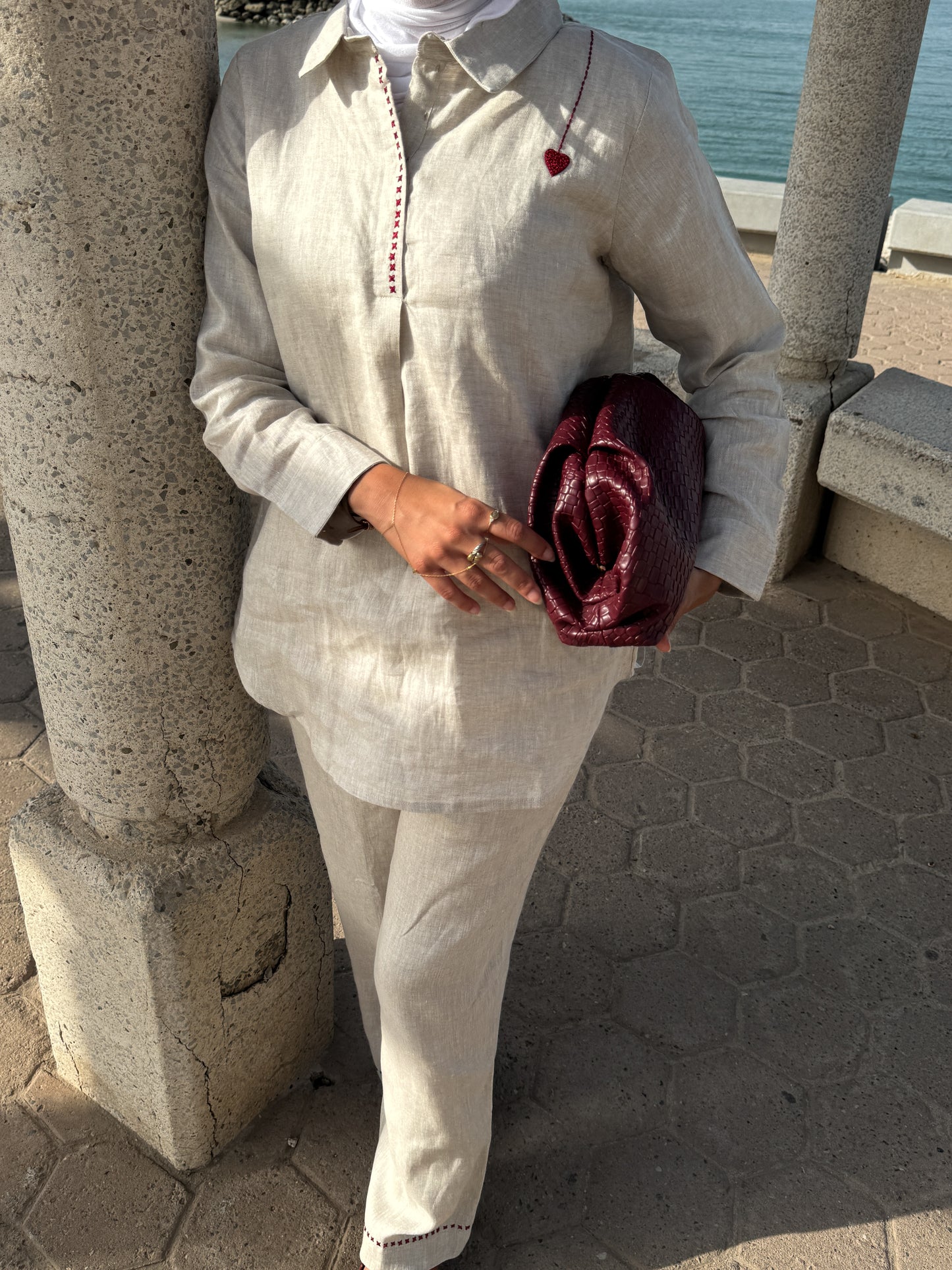 Person wearing a light-colored outfit with a red clutch, standing near stone columns with a body of water in the background.