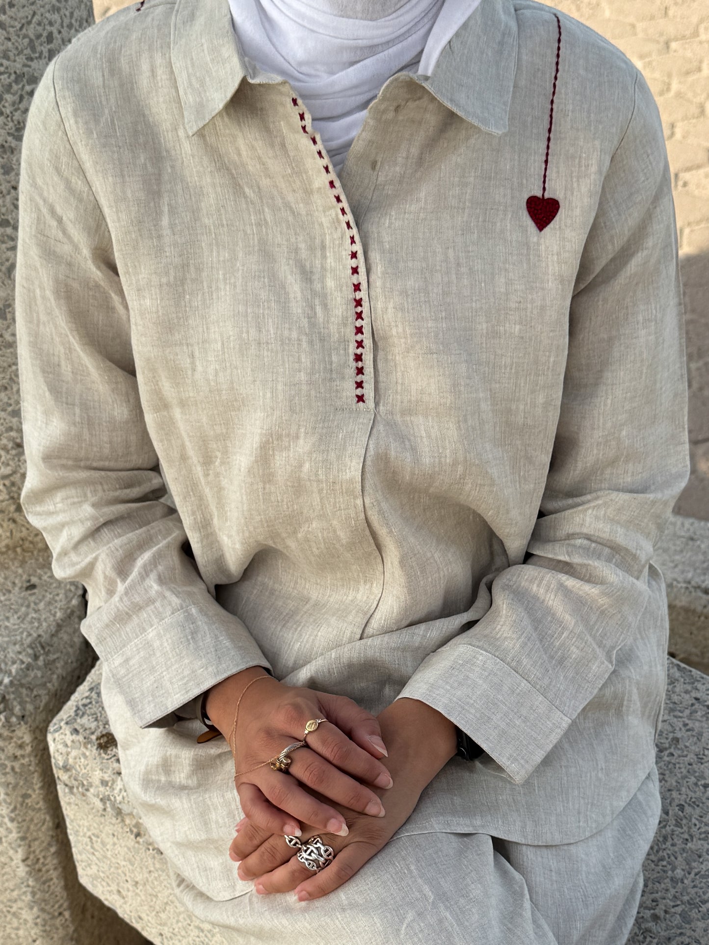 Person wearing a light-colored shirt with decorative stitching sitting on a stone surface.