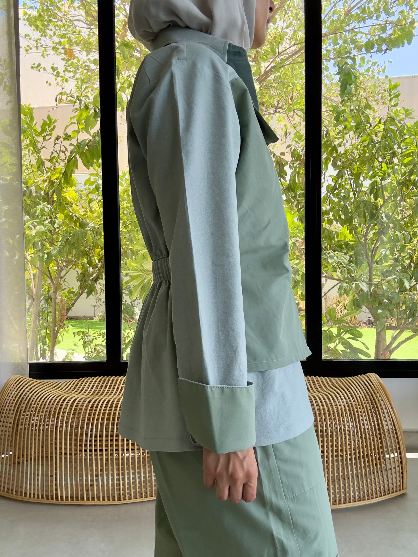 Person wearing a light green outfit standing in front of a window with a view of trees.