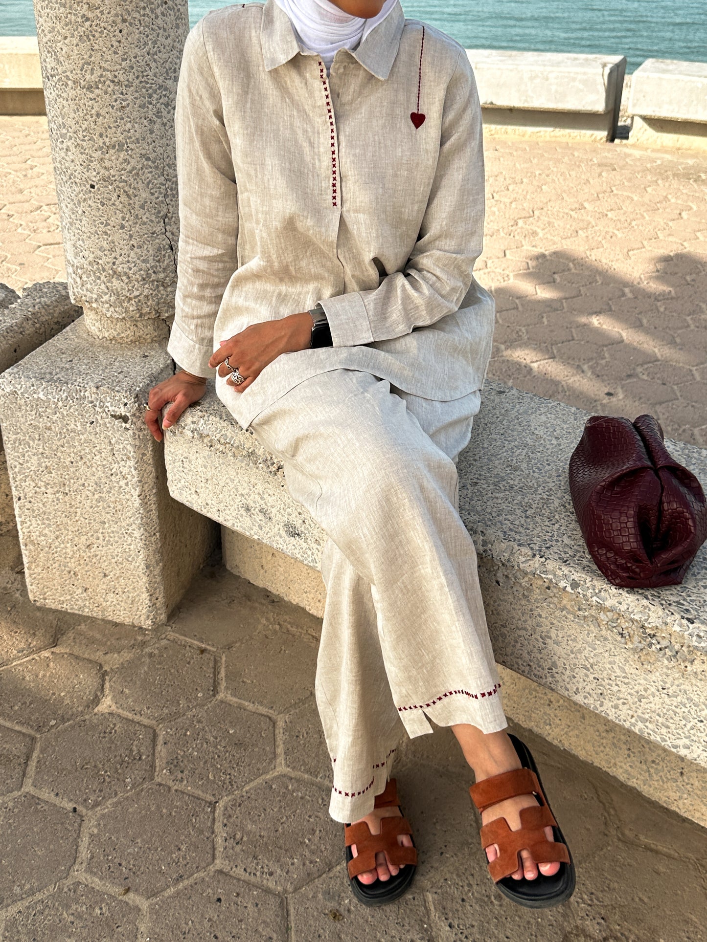 Person sitting on a stone bench by the sea wearing a light-colored outfit with brown sandals.