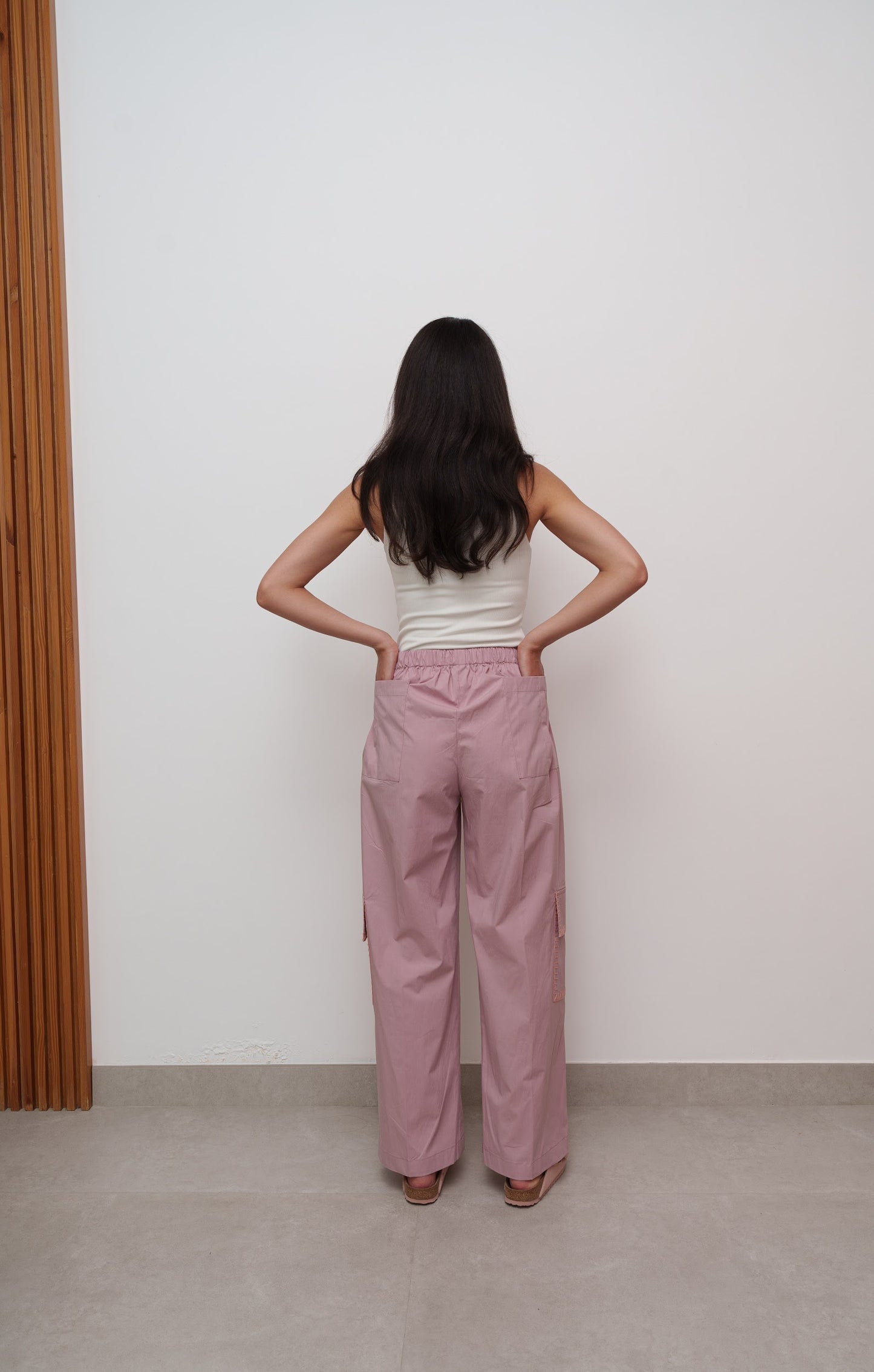 Person wearing a white top and pink pants standing against a plain wall.