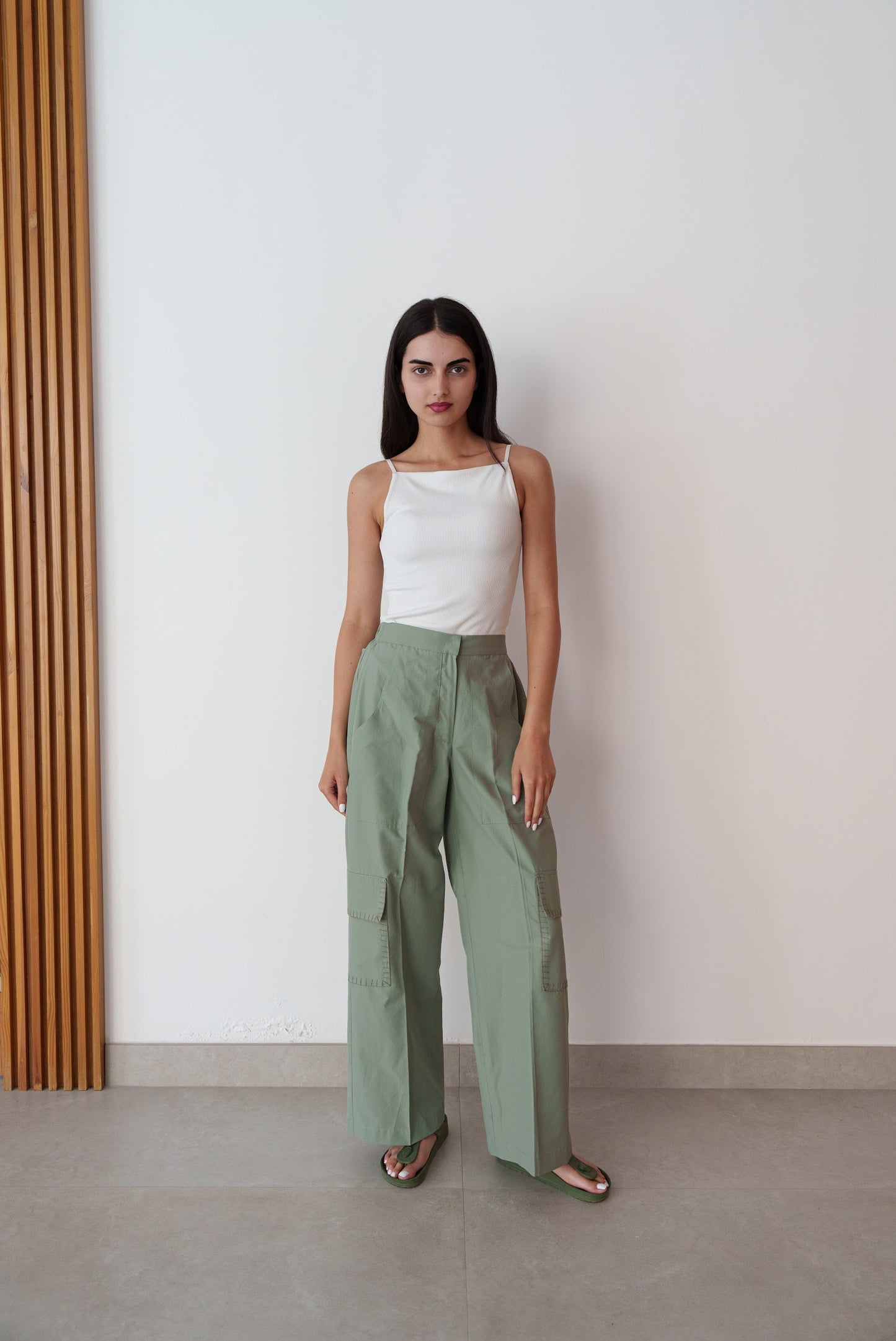 Woman wearing a white top and green pants standing against a plain wall.
