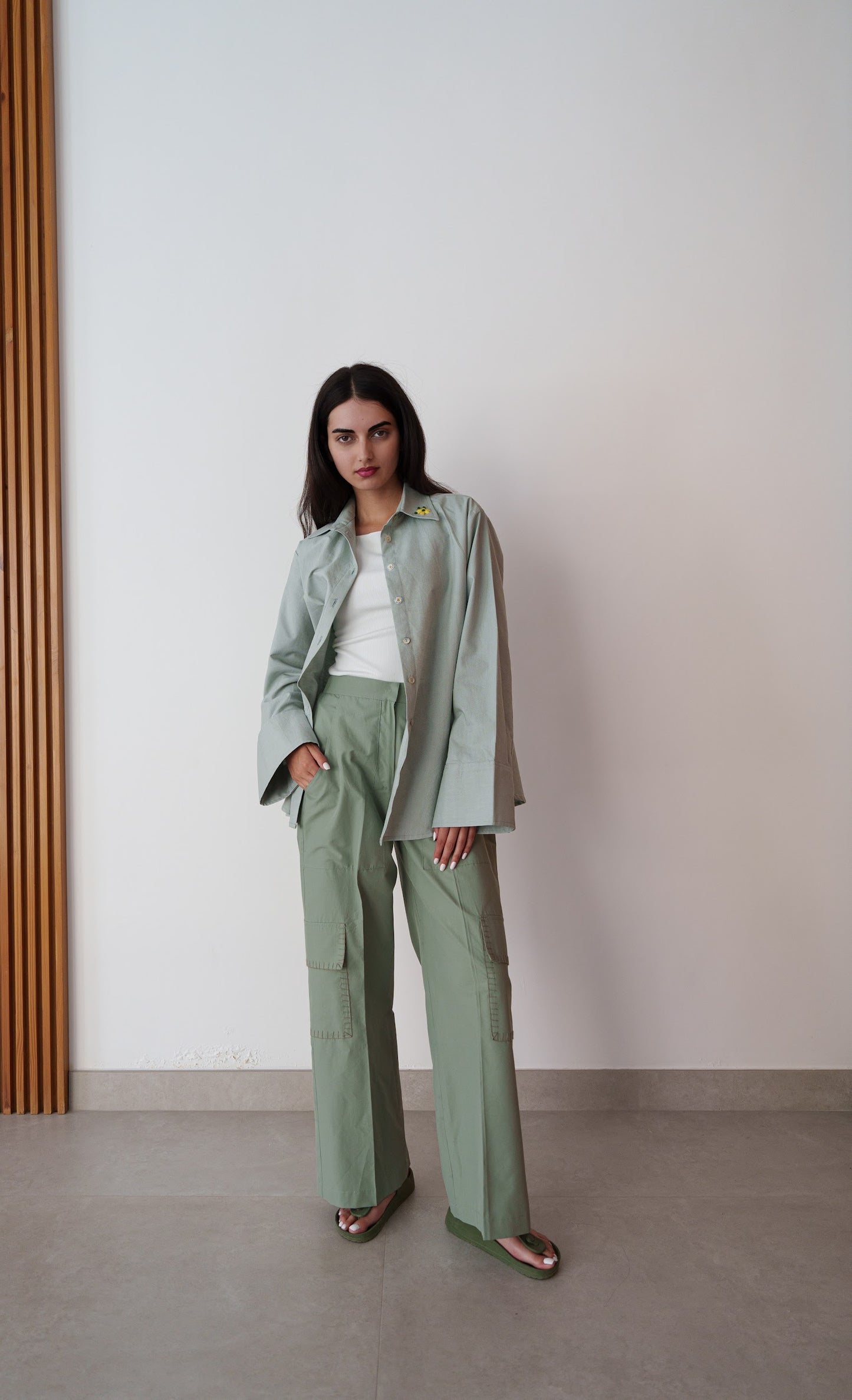 Woman wearing a light green jacket and pants standing against a plain wall.