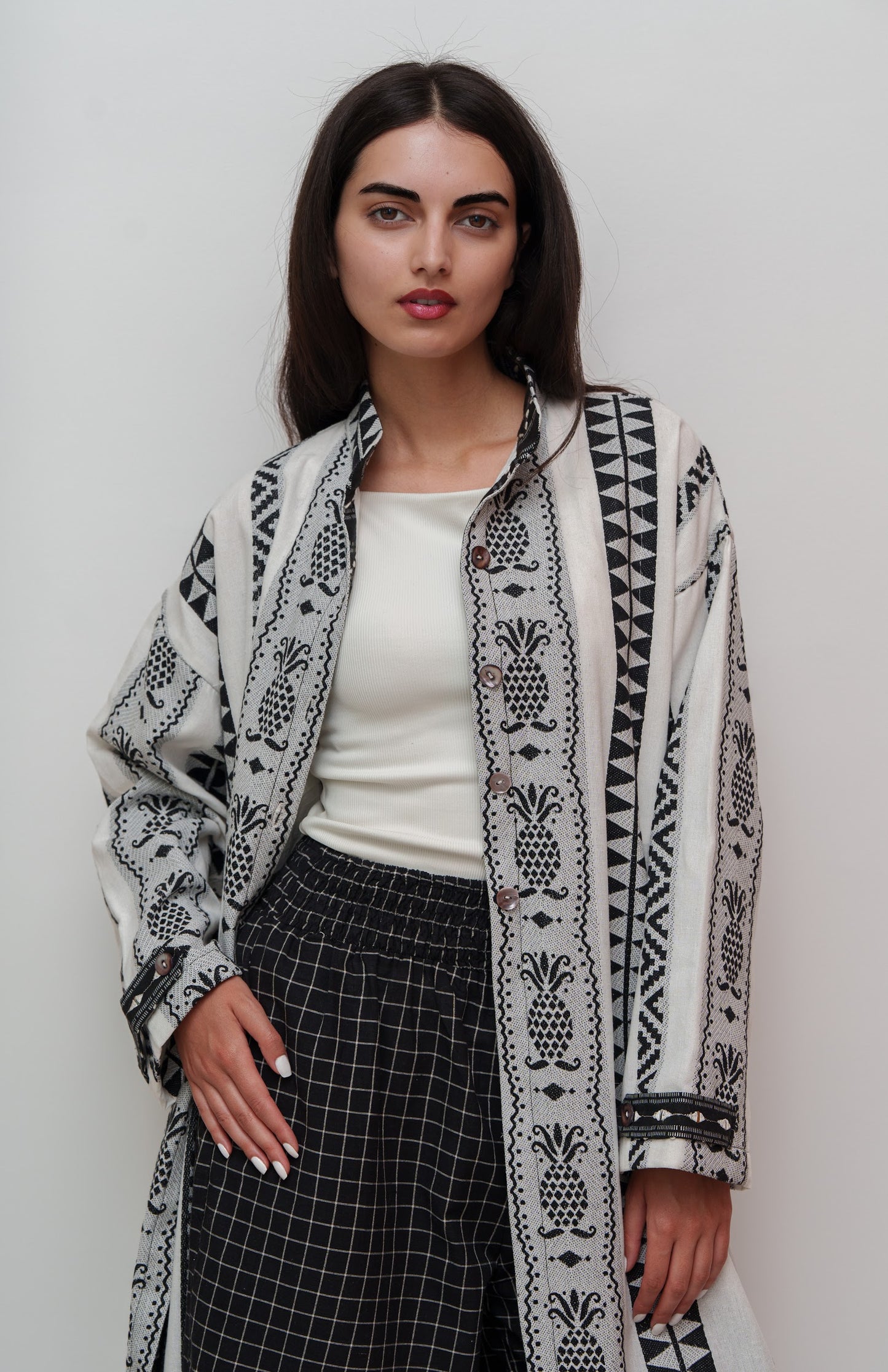 Woman wearing a long, patterned jacket over a white top and black pants on a plain background