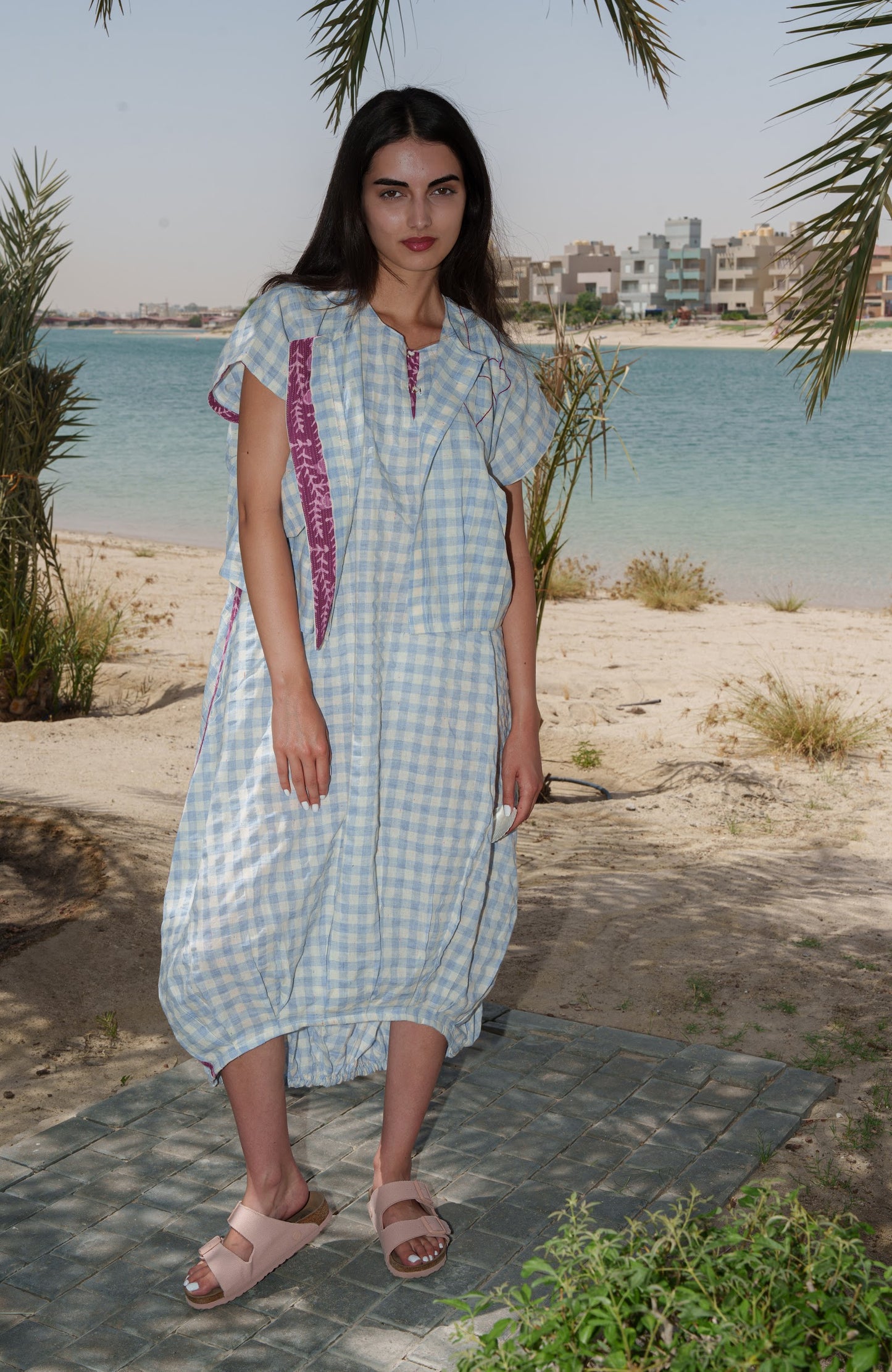 Woman in a checkered dress standing on a sandy beach with palm trees and water in the background