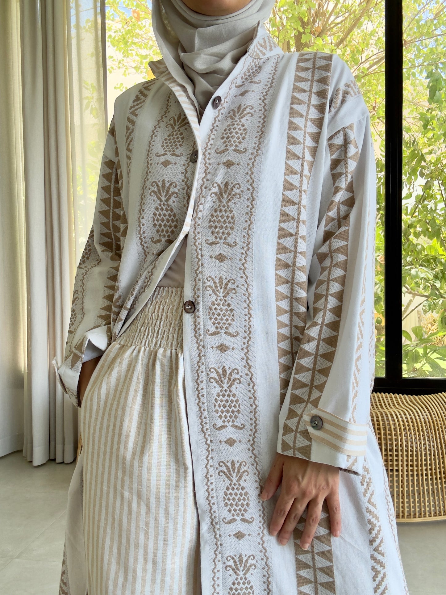 Woman in white embroidered outfit with brown detailing, posing near large windows in soft indoor light.