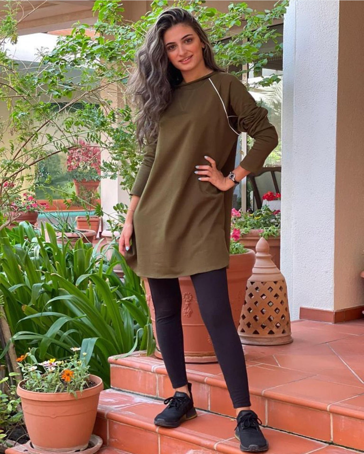 A person wearing a green khaki active top paired with black leggings, standing near potted plants.