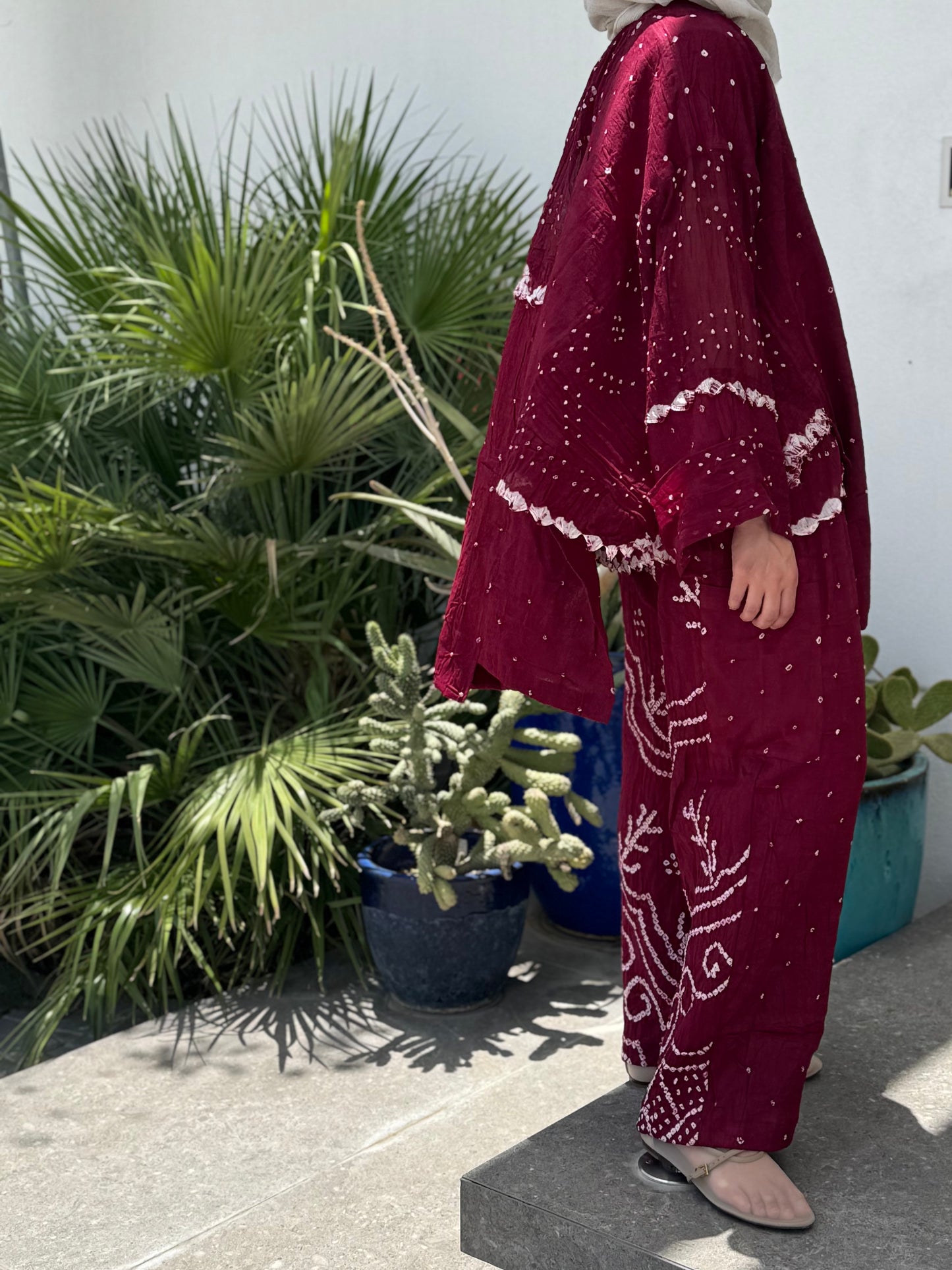 Person wearing a burgundy outfit with white patterns standing outdoors near plants.