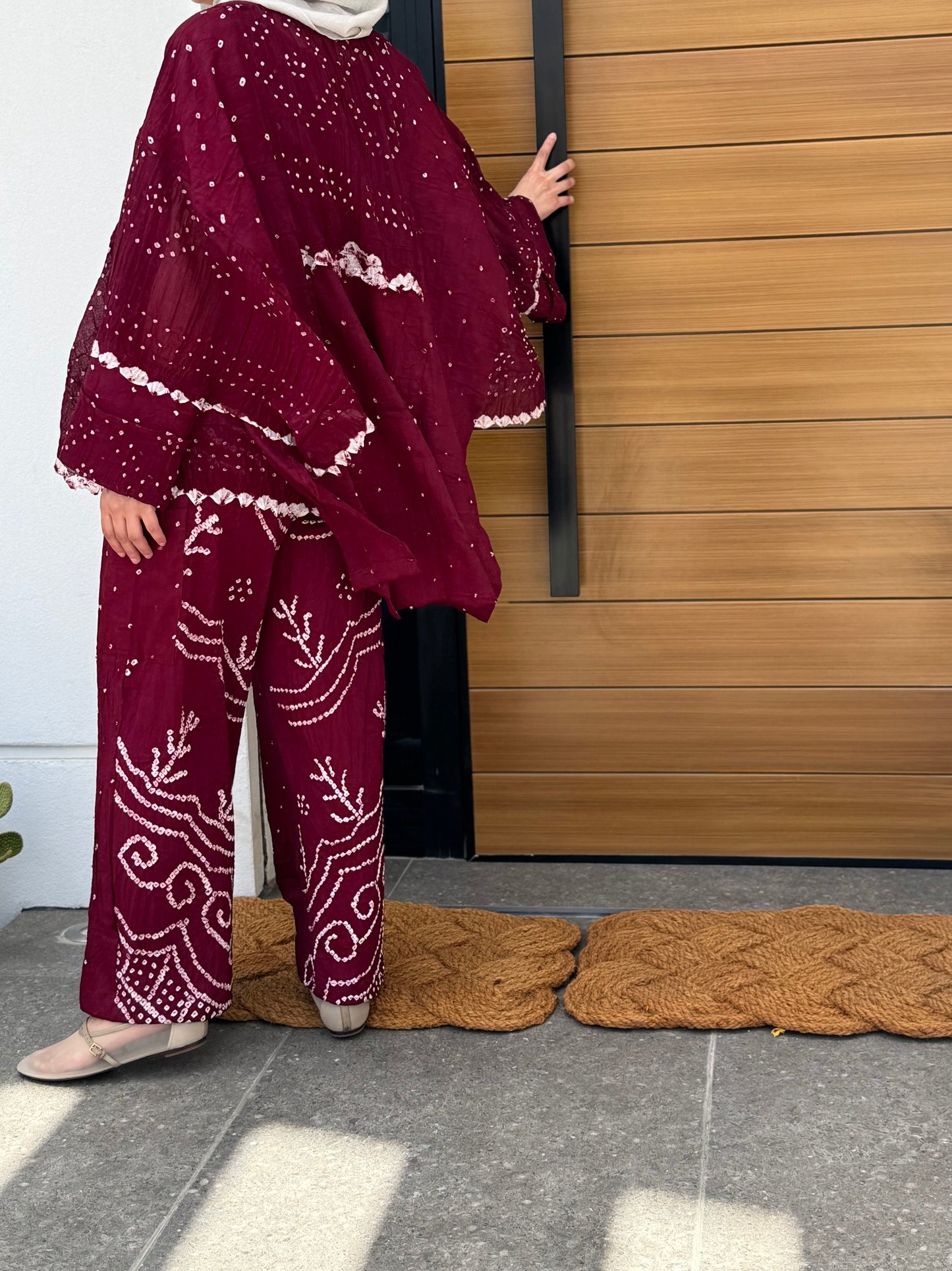 Person wearing a burgundy outfit with white patterns standing next to a wooden door.