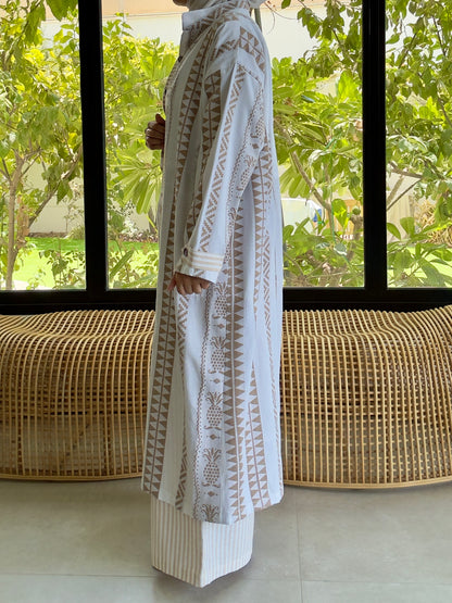 Model in white linen outfit with detailed embroidery, indoors near large windows with greenery outside