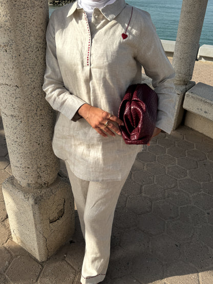 Person wearing a light-colored outfit with a red clutch, standing near stone columns with a body of water in the background.