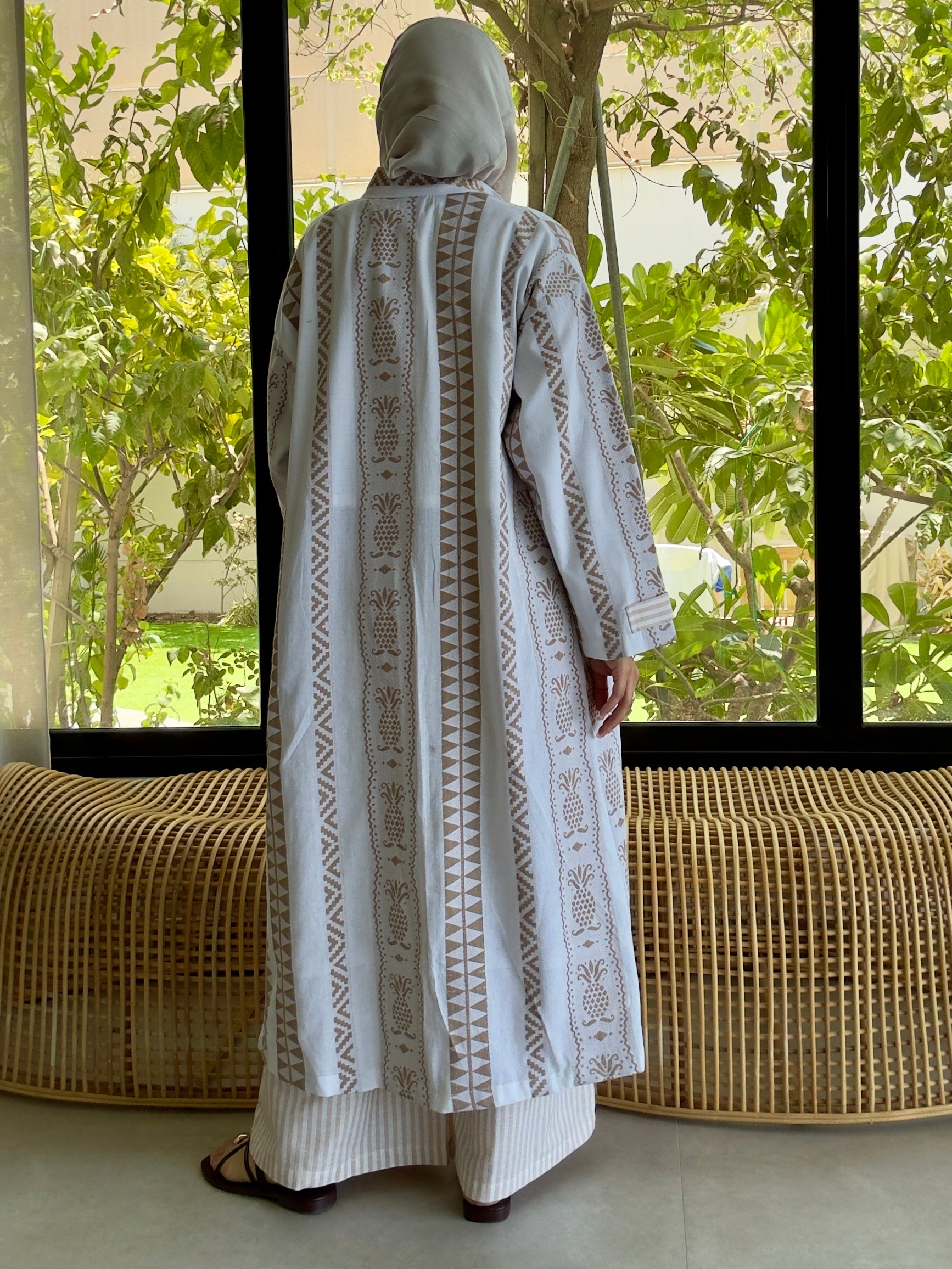 Back view of white linen dress with delicate embroidery, modeled in front of glass doors with greenery outside.
