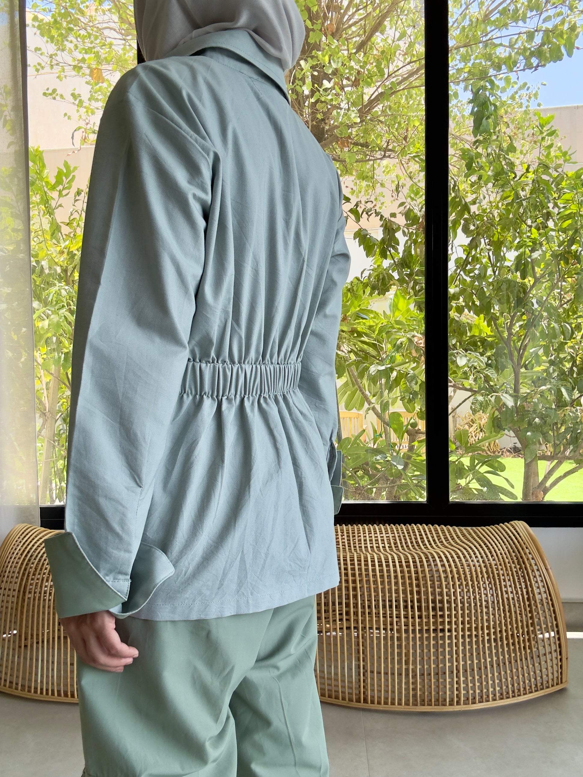Person wearing a light green outfit standing in front of a window with greenery outside.