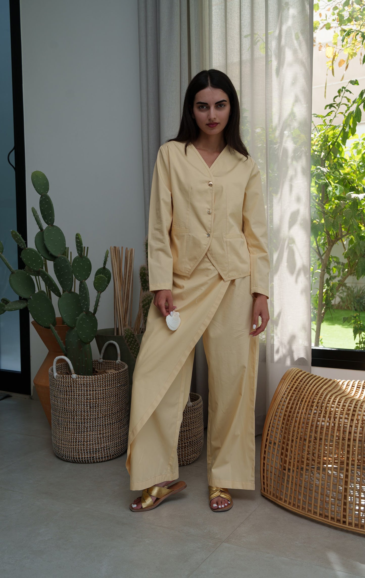 Woman in a beige outfit standing in a room with plants and a window.