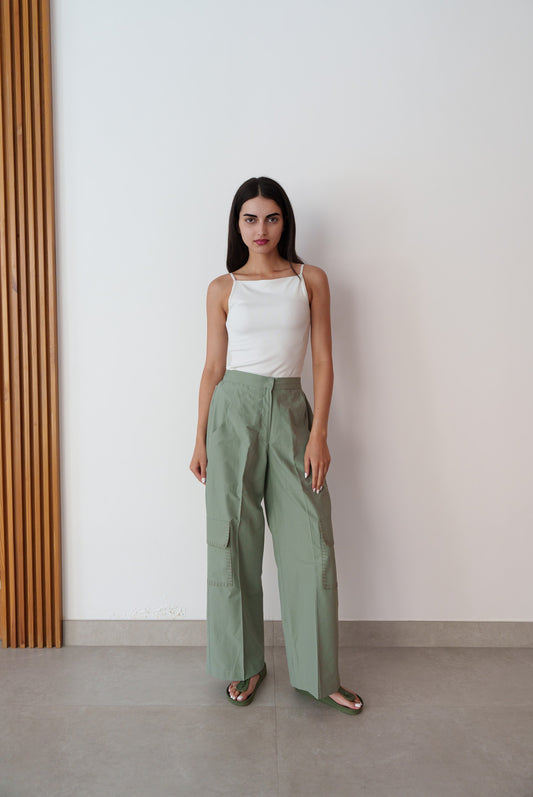 Woman wearing a white top and green pants standing against a plain wall.