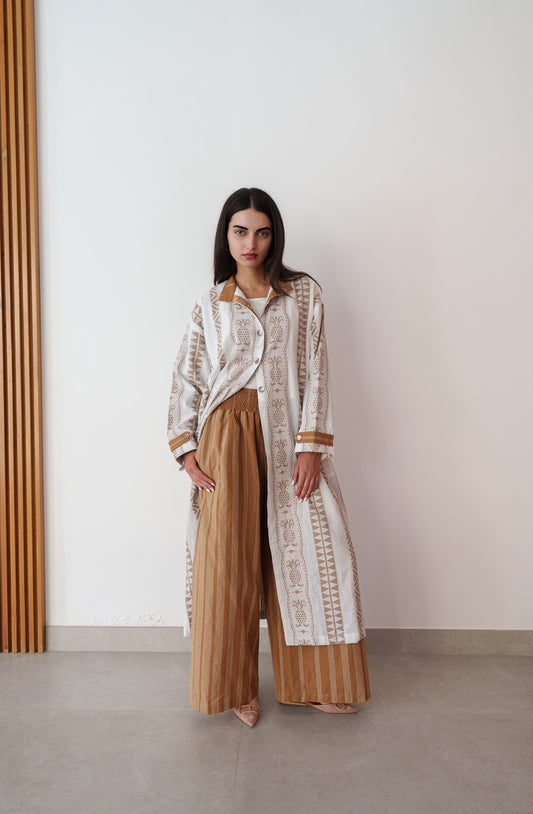 Woman wearing a long, patterned coat over a white shirt and brown pants against a plain wall.