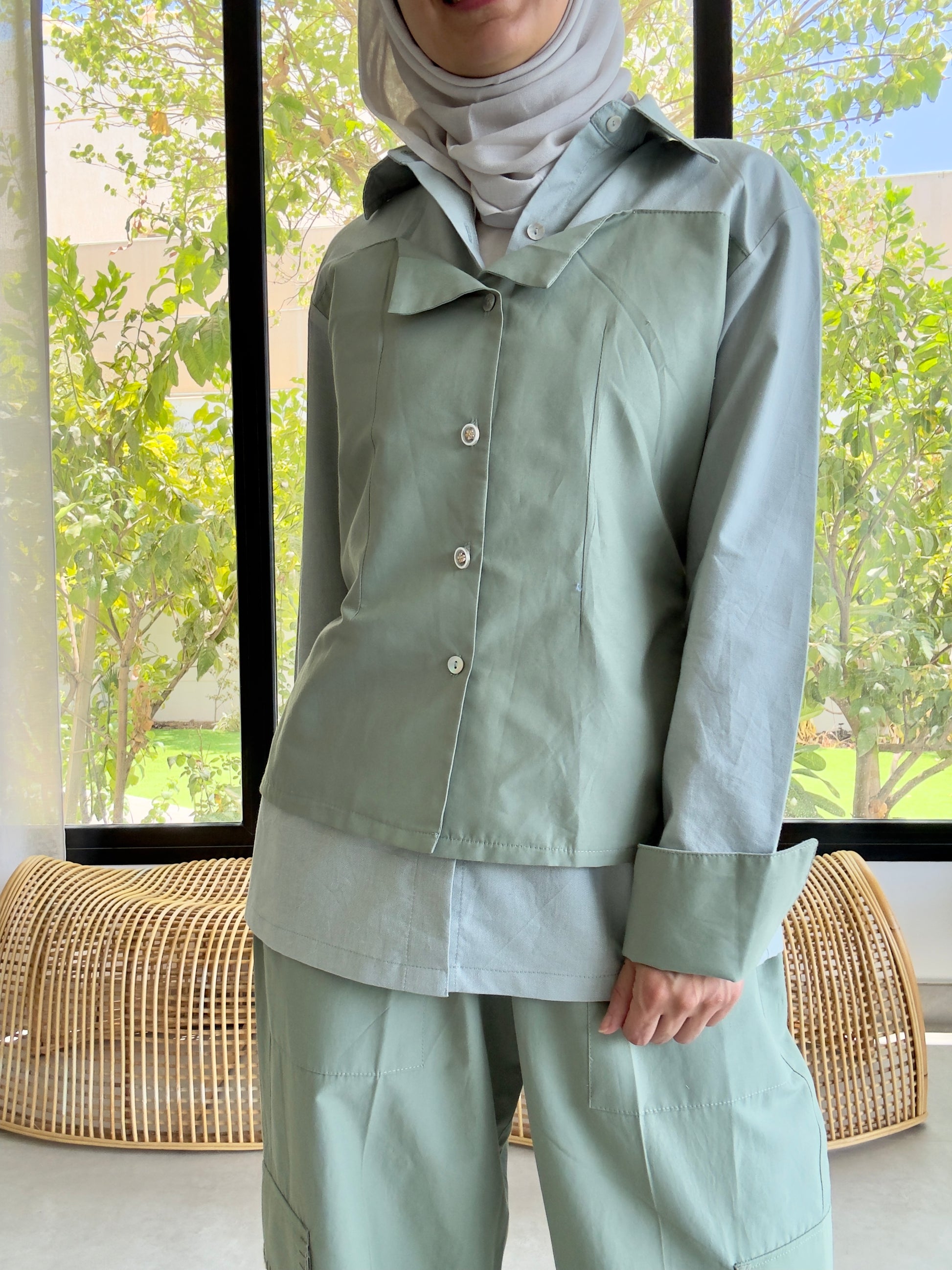 Person wearing a light green outfit with a hijab in a room with large windows.