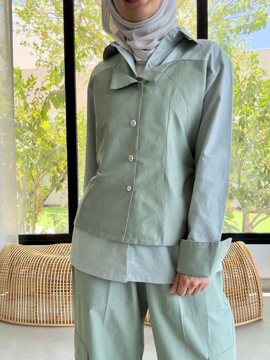 Person wearing a light green outfit with a hijab in a room with large windows.