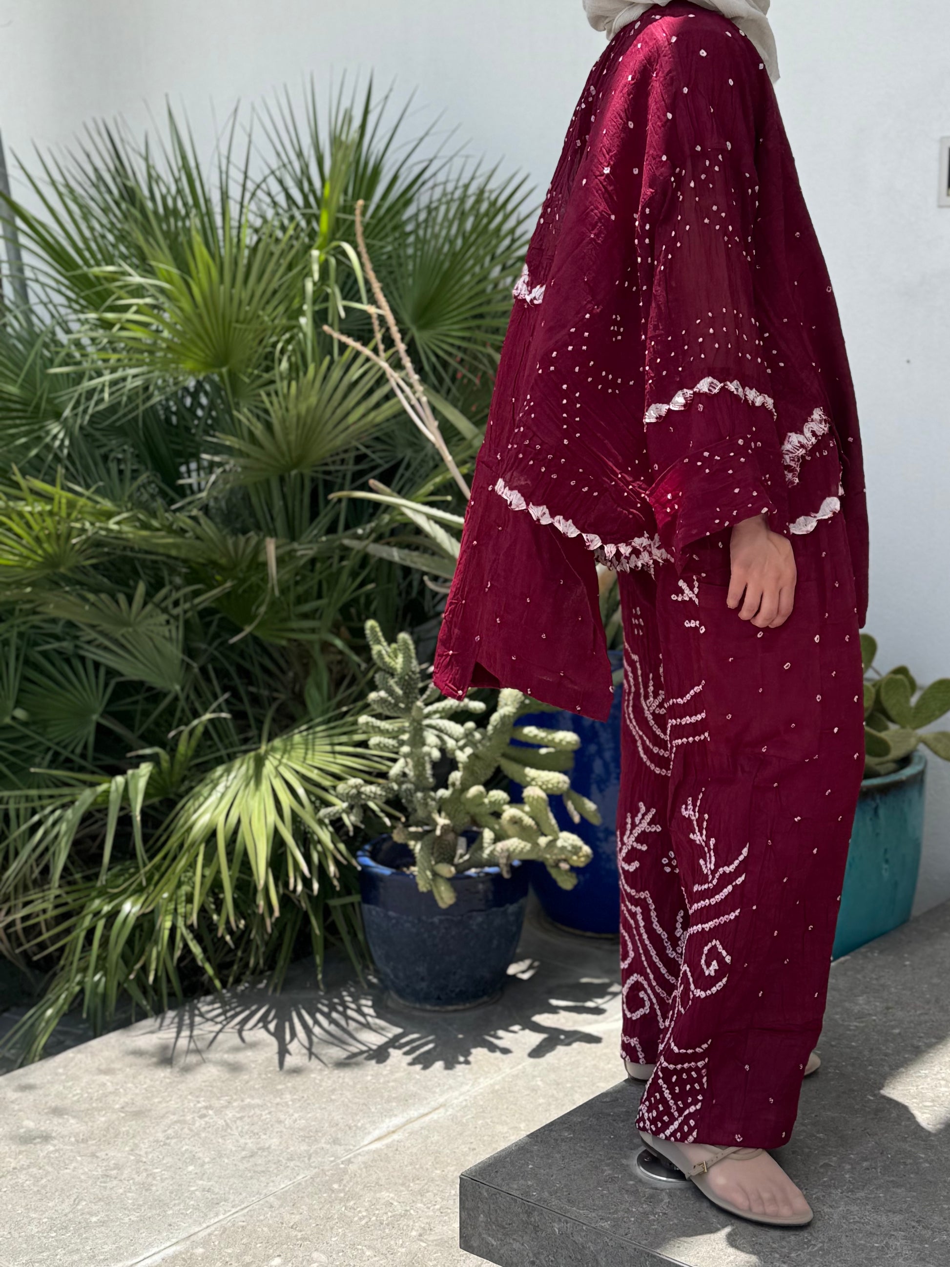 Person wearing a burgundy outfit with white patterns standing outdoors near plants.